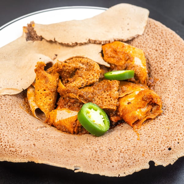 Spicy Ethiopian Doro Wat chicken stew with injera bread and sliced jalapeños on a white plate.