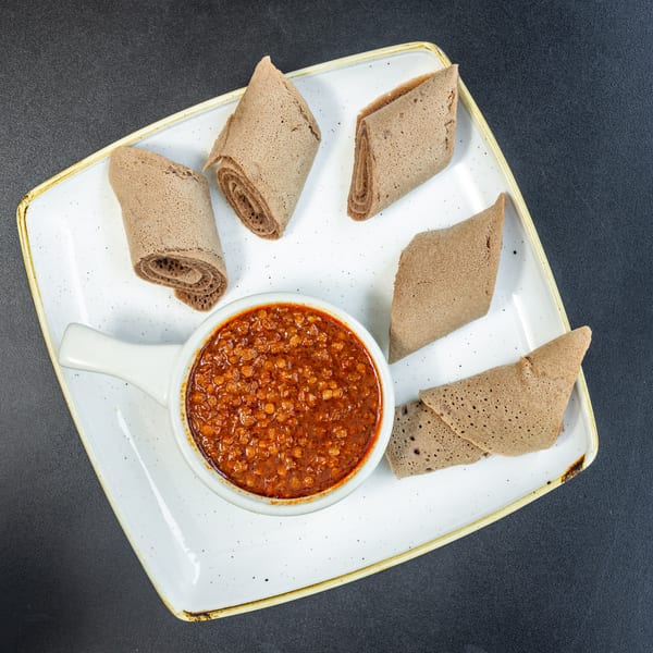A white rectangular plate holds a bowl of red lentil stew surrounded by injera bread pieces.