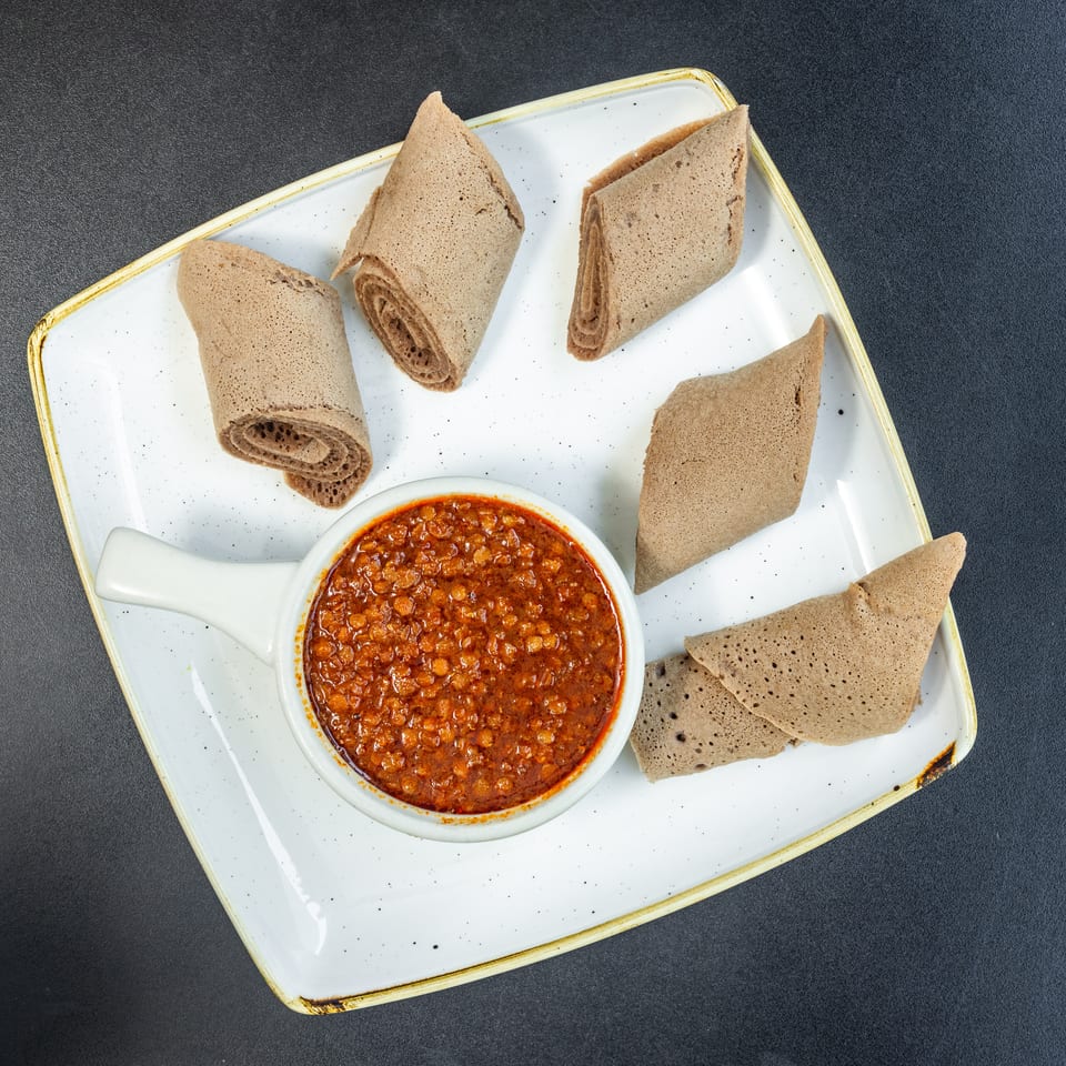 A white rectangular plate holds a bowl of red lentil stew surrounded by injera bread pieces.