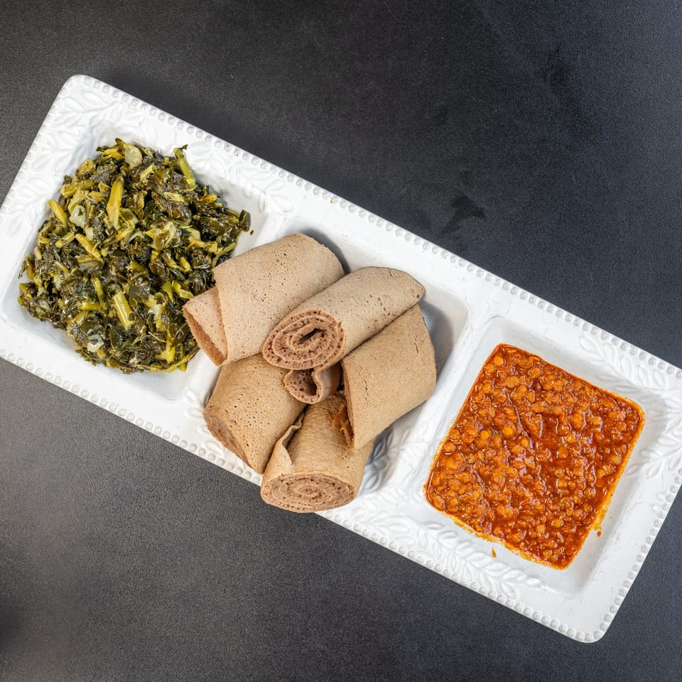 A white rectangular plate with three sections, holding green collard greens, rolled injera bread, and red lentils.