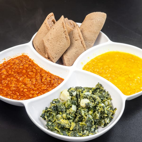 A white divided dish holds injera bread, red lentils, yellow split peas, and collard greens.