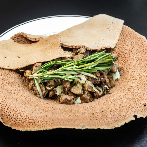 Ethiopian injera bread with meat, onions, and rosemary on a white plate.