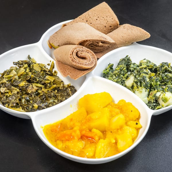 A white segmented dish holds injera bread, collard greens, spinach, and potato stew on a dark surface.