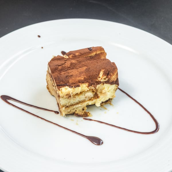 A slice of tiramisu with cocoa powder and chocolate sauce on a white plate.