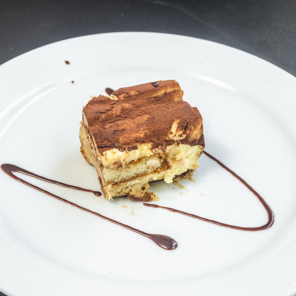 A slice of tiramisu with cocoa powder and chocolate sauce on a white plate.