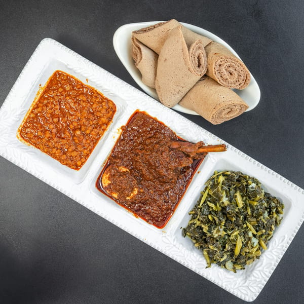 A white rectangular platter with three Ethiopian dishes: lentils, chicken stew, and collard greens, with injera bread.